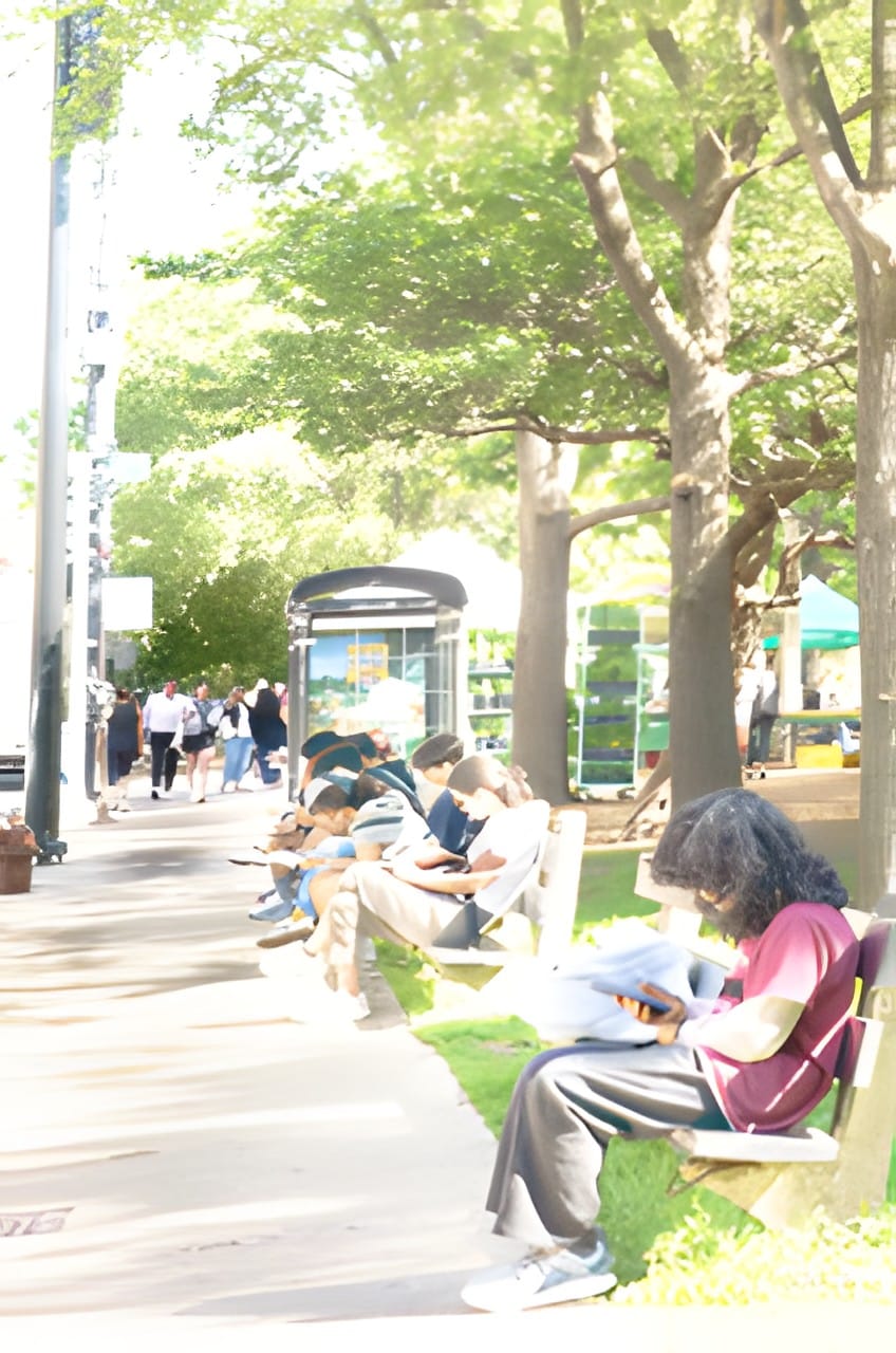 People sitting under leafy trees beside a city sidewalk, soft green