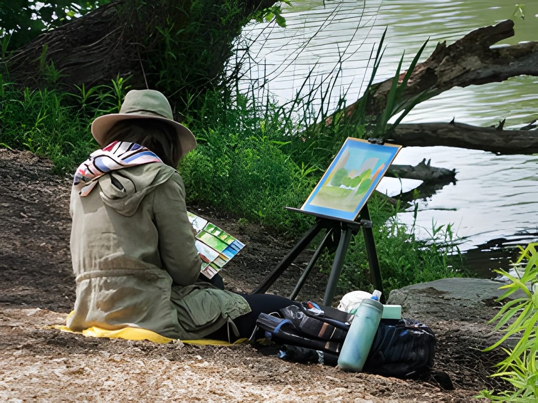 A woman painting at an easel outdoors beneath a large tree